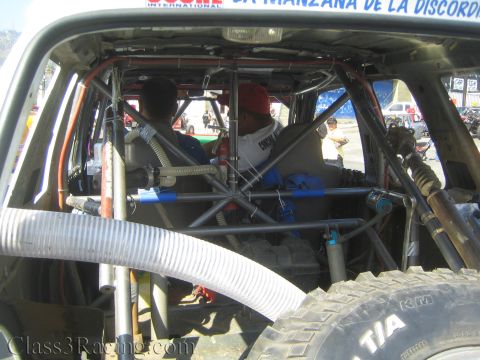 1990 Ford Bronco - View into an off-road vehicle's caged interior with two people seated, a corrugated air hose and tires in the foreground.