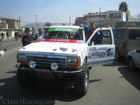 1990 Ford Bronco - white lifted Ford race truck marked 30 sits on a street with its door open and several people gathered around.