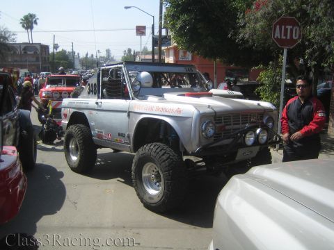 1990 Ford Bronco - silver early Ford Bronco-style off-road truck marked 348 parked on a street beside a stop (ALTO) sign and onlookers.