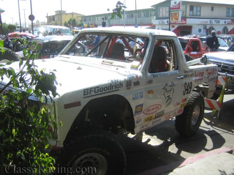 1990 Ford Bronco - white off-road race truck marked 349 covered in BFGoodrich and sponsor decals parked among other trucks at a staging lot.