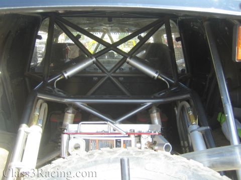 1990 Ford Bronco - Looking into the rear of an off-road vehicle at the tubular roll cage X-bracing, coilover shocks and a mounted spare tire.