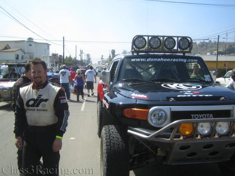 1990 Ford Bronco - smiling man in an Orange Racing fire suit poses next to a black Toyota FJ Cruiser off-road race truck with a roof light bar on a town stree…