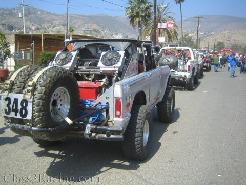 1990 Ford Bronco - Rear three-quarter view of a silver number 348 early Ford Bronco off-road racer with exposed roll cage and rear-mounted spare, lined up wit…