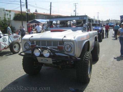 1990 Ford Bronco - Front view of a silver early Ford Bronco off-road racer with a tube bumper, round headlights and a Ford-badged grille, parked among other r…