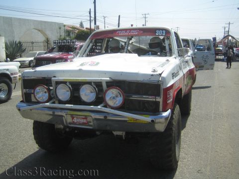 1990 Ford Bronco - Front view of a white-and-red number 303 off-road race truck with a chrome bumper, four round driving lights and Phils Inc sponsor letterin…