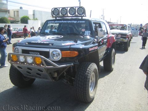 1990 Ford Bronco - Front three-quarter view of a black Toyota FJ Cruiser off-road racer, number 302, with a roof light bar and tube bumper, parked with other…
