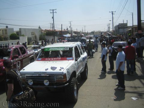 1990 Ford Bronco - white number 30 off-road race truck staged on a crowded street lined with spectators and other race vehicles under power lines.