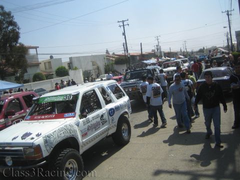 1990 Ford Bronco - White number 30 SUV off-road racer parked among other race trucks as a crowd walks down a sunlit dirt street.