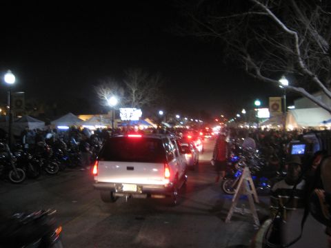 1986 Ford Bronco - A busy street scene at night with a white SUV in the foreground, surrounded by motorcycles and a crowd of people.