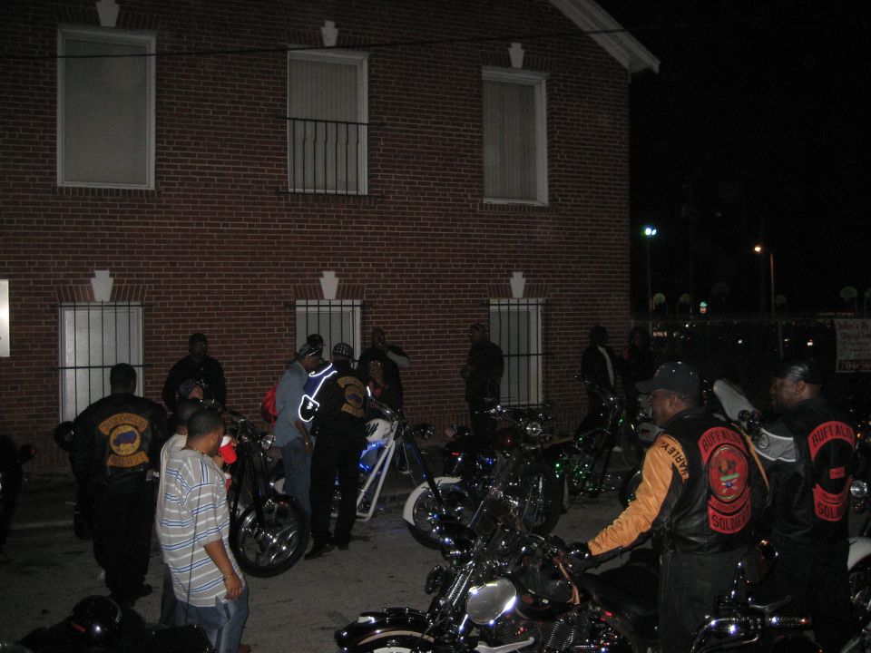 Motorcycles parked outside a brick building at night, with people gathered around