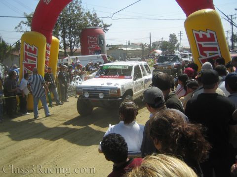 1990 Ford Bronco - White number 30 SUV off-road racer drives under a red Tecate inflatable arch while crowds line both sides of the dirt course.