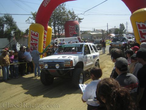 1990 Ford Bronco - White number 30 SUV off-road racer passes under the red Tecate start arch on a dirt course, framed by spectators and palm trees.