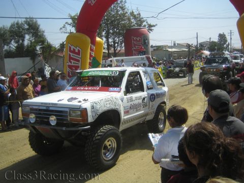 1990 Ford Bronco - Side view of a heavily decaled white number 30 SUV off-road racer driving along a dusty course lined with spectators and Tecate banners.