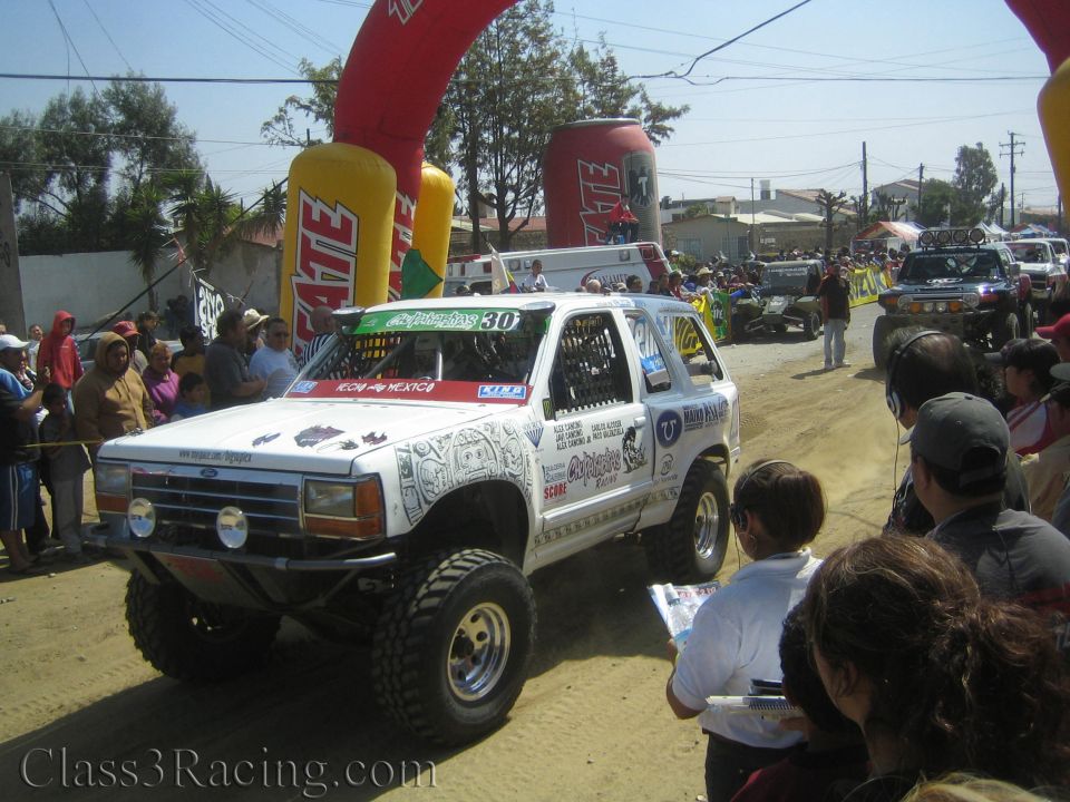 White SUV racer number 30 covered in sponsor decals driving along a dusty spectator course