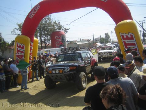 1990 Ford Bronco - Black Toyota FJ Cruiser off-road racer rolls forward under the red Tecate start arch with a red truck behind and a dense crowd watching.