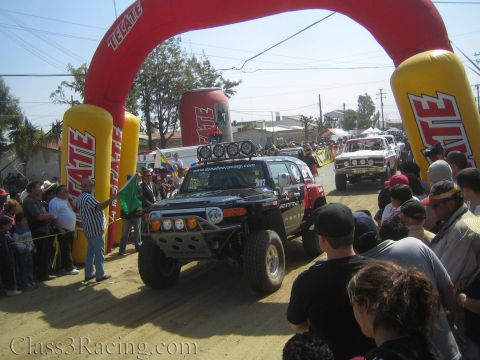 1990 Ford Bronco - black Toyota FJ Cruiser off-road racer with four roof lights crosses under the red Tecate arch as a green flag waves and crowds watch.