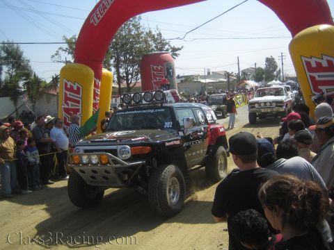1990 Ford Bronco - black off-road racing Toyota FJ Cruiser numbered 302 drives under a red Tecate inflatable arch as spectators line the dusty course.