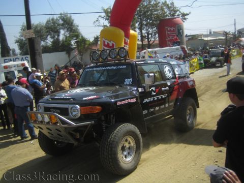 1990 Ford Bronco - black Toyota FJ Cruiser race truck #302 with Nitto and Donahoe Racing decals turns on a dusty street past a crowd.
