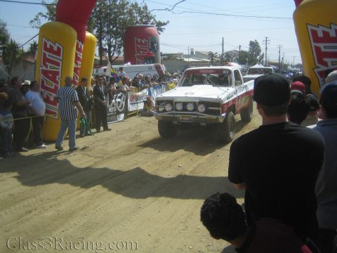 1990 Ford Bronco - white vintage pickup-style off-road race truck heads toward a red Tecate arch as spectators watch from both sides.