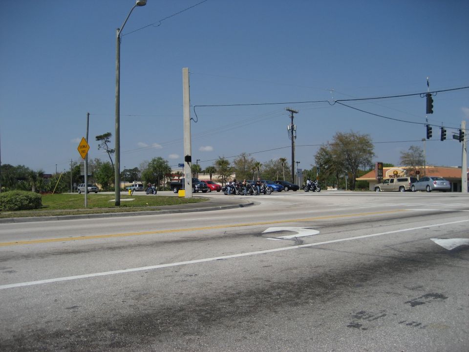 Several motorcycles are stopped at a traffic light intersection with cars and buildings in the background
