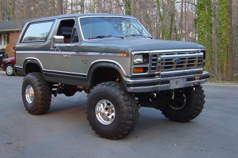 1986 Ford Bronco - A two-tone gray and silver 1986 Ford Bronco full-size SUV shown from a front three-quarter angle, heavily lifted on large mud-terrain tires…