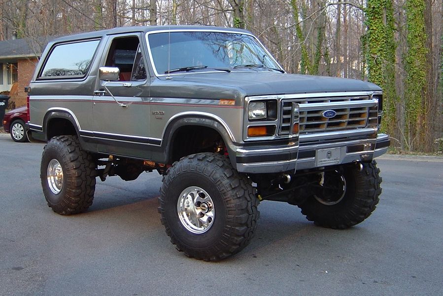 Lifted gray Ford Bronco SUV on big mud tires, front 3/4 view in a wooded driveway.