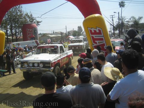 1990 Ford Bronco - white-and-red vintage off-road race truck passes under inflatable Tecate arches while a dense crowd looks on.