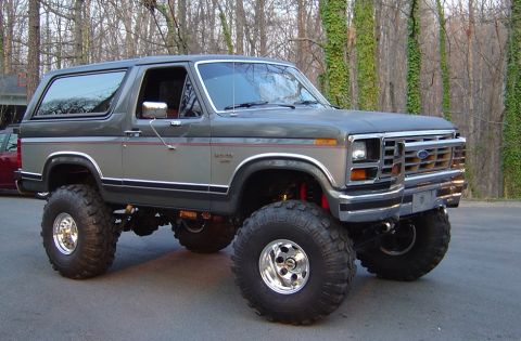 1986 Ford Bronco - A two-tone gray lifted Ford Bronco full-size SUV shown from the front three-quarter angle, riding on large off-road mud tires and polished…