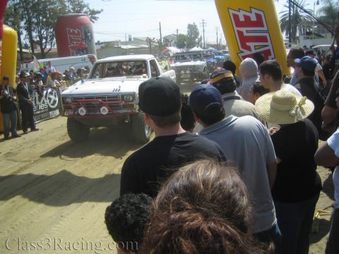 1990 Ford Bronco - white off-road race truck #349 with round driving lights passes under a Tecate arch among a tight crowd.
