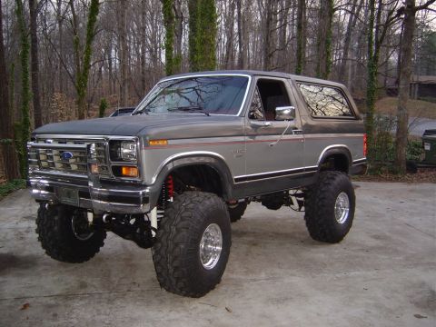 1986 Ford Bronco - A two-tone gray and silver 1980s Ford Bronco full-size SUV heavily lifted on large mud-terrain tires with polished aluminum wheels, chrome…