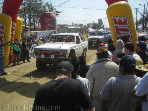 1990 Ford Bronco - white off-road race truck #349 drives between yellow Tecate arches as a crowd of spectators lines the dusty course.