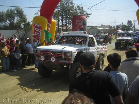 1990 Ford Bronco - white vintage Ford-style off-road race truck #349 with red tube bumper and BFGoodrich livery drives past a flagger and crowd.