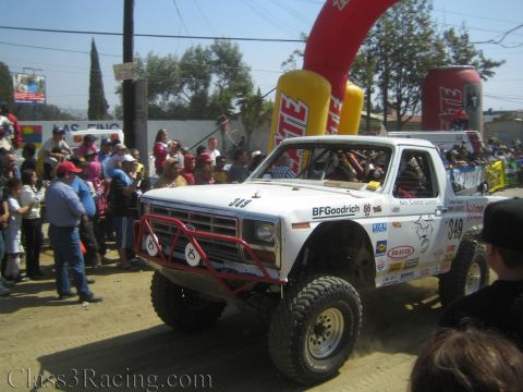 1990 Ford Bronco - white vintage Ford-style off-road race truck #349 with red tube bumper and BFGoodrich decals passes under a Tecate arch.