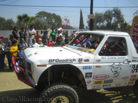 1990 Ford Bronco - white off-road race truck numbered 34 with BFGoodrich and Deaver decals idles past a crowd of spectators on a dirt course.