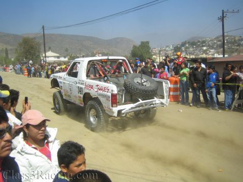 1990 Ford Bronco - Rear view of white off-road race truck #349 lettered Anger Issues with spare tire, driving down a dusty road past spectators.