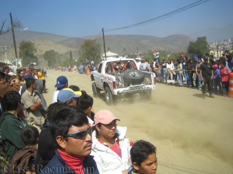 1990 Ford Bronco - white off-road race truck drives down a dusty road flanked by a large crowd, with bare hills in the distance.