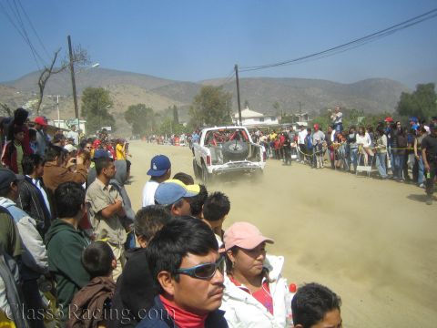 1990 Ford Bronco - distant white off-road race truck moves along a wide dusty desert road as spectators crowd the foreground.