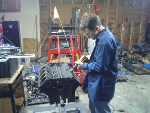 1976 Ford F-100 - A person is using a tool to work on an engine block in a cluttered garage setting.