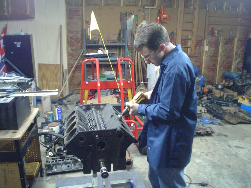 Man in blue shirt works on a black engine block on a stand in a garage