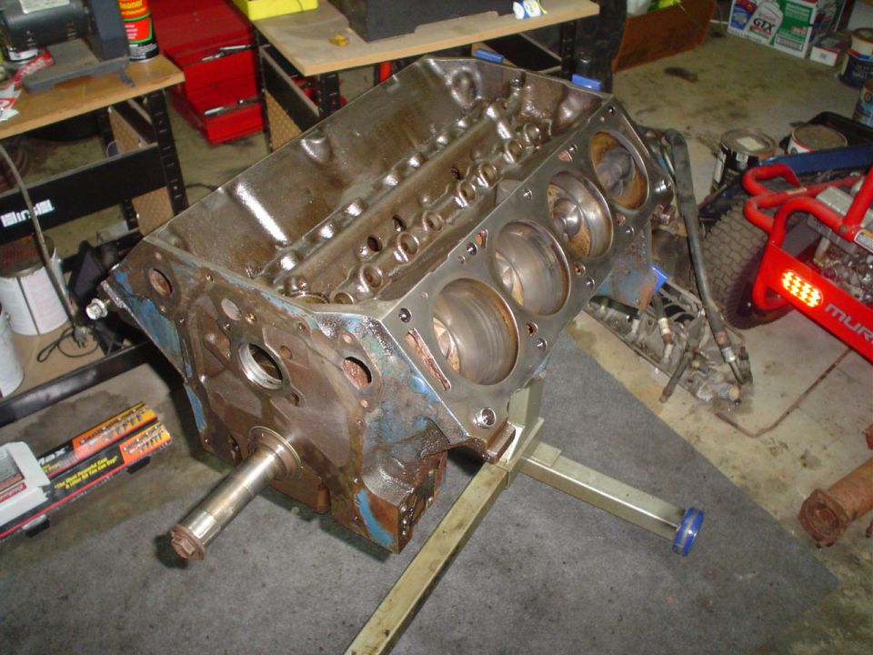 Engine block on a stand, showing cylinder bores and crankshaft snout. Rusty and blue paint visible