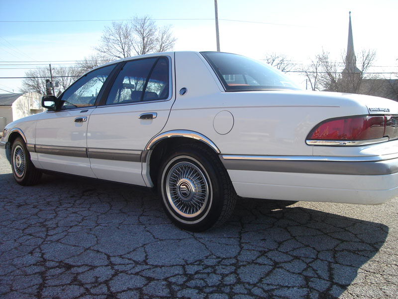 White Mercury Grand Marquis parked on cracked asphalt. Church steeple visible in background