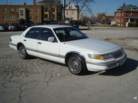 1992 Ford Crown Victoria - A white sedan, likely from the 1990s, is parked on a cracked asphalt lot.