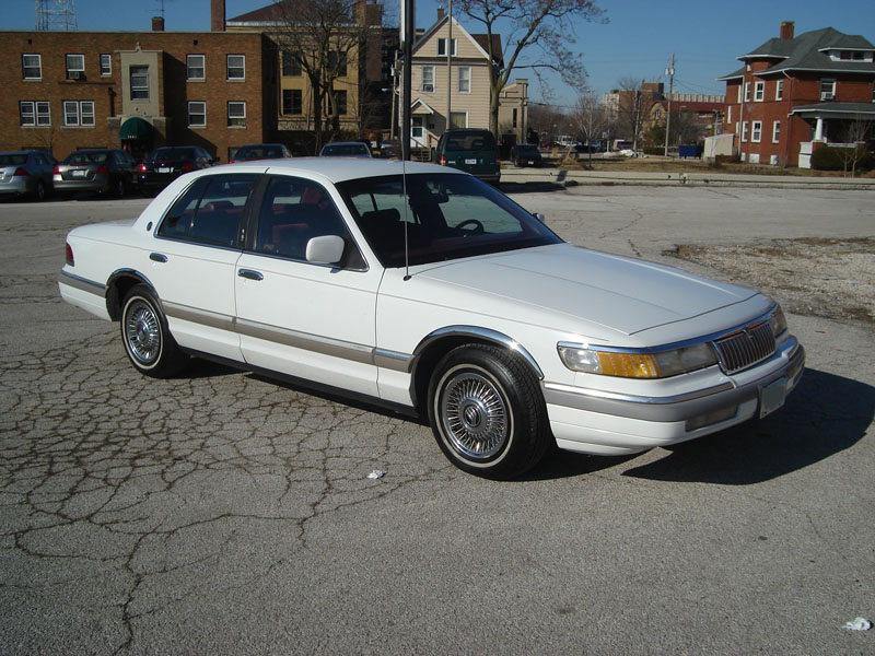 White 1990s sedan with chrome trim and wire wheels parked on cracked asphalt