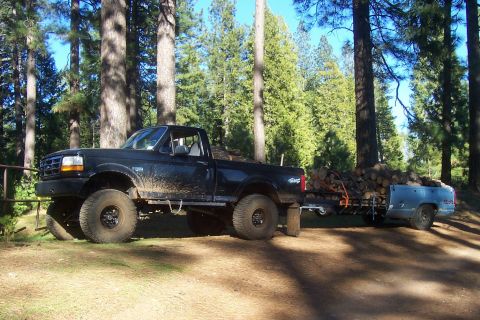 1994 Ford F-150 - A lifted black Ford pickup truck tows a trailer full of firewood through a wooded area.