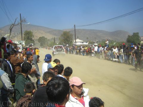 1990 Ford Bronco - small off-road race vehicle drives down a dusty dirt street as crowds of spectators line both sides below arid hills.