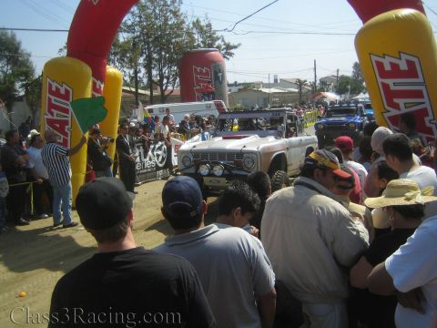 1990 Ford Bronco - silver early Ford Bronco race truck waits under Tecate inflatable arches as spectators watch from the foreground.
