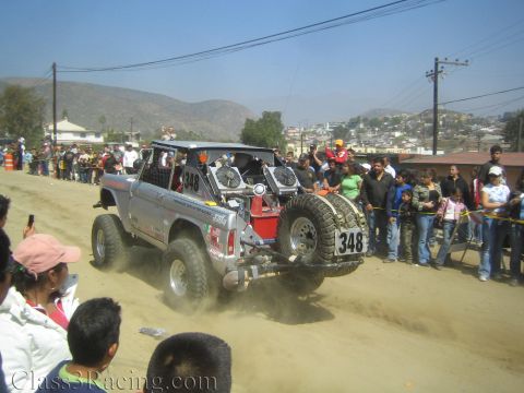 1990 Ford Bronco - Rear view of silver off-road race truck number 348 with a roof light bar and mounted spare tire on a dusty road.