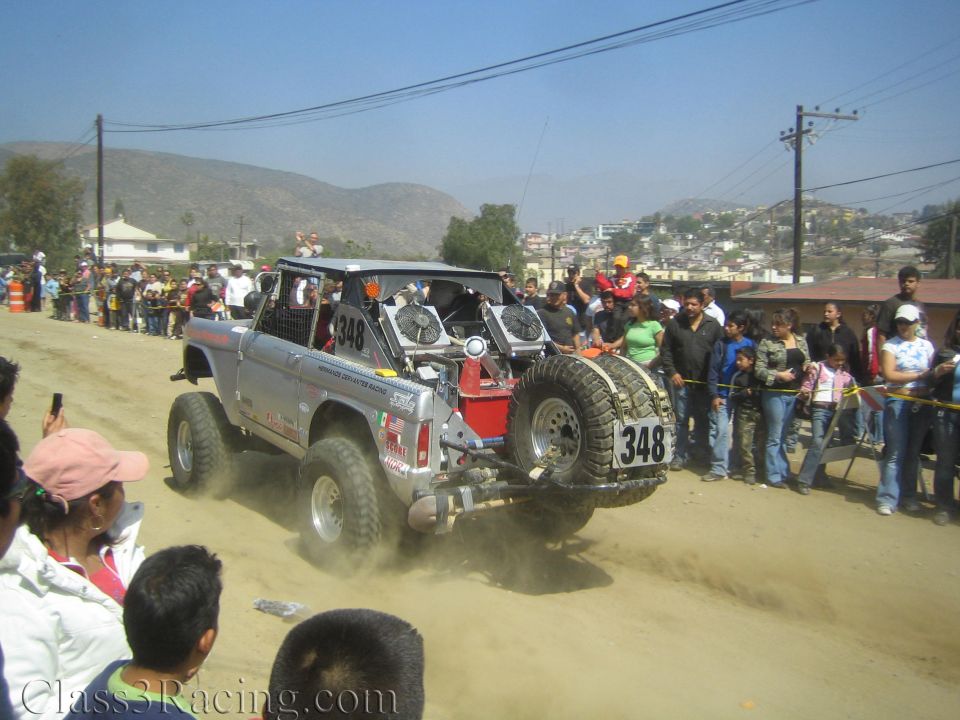 Rear of silver race truck 348 with light bar and spare tire kicking up dust