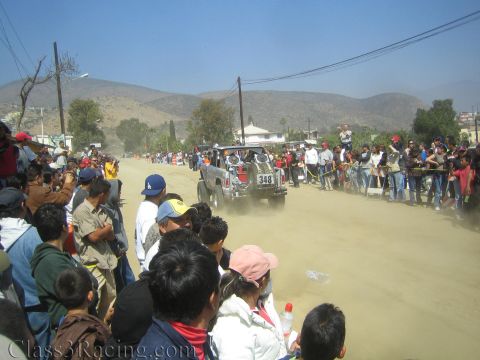 1990 Ford Bronco - Off-road race truck number 348 heads down a dusty dirt road flanked by spectators with hills in the background.