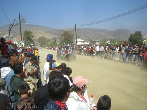 1990 Ford Bronco - distant off-road race truck drives down a dusty street lined with spectators beneath dry hills.